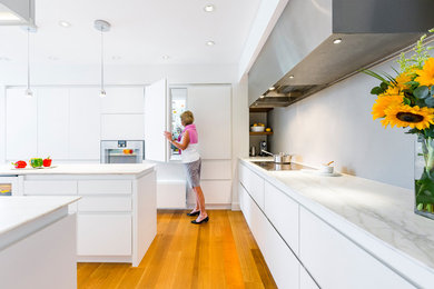 Mid-sized modern u-shaped eat-in kitchen in Cincinnati with a single-bowl sink, flat-panel cabinets, white cabinets, marble benchtops, grey splashback, marble splashback, stainless steel appliances, medium hardwood floors, with island and brown floor.