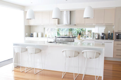 This is an example of a large contemporary galley eat-in kitchen in Sydney with an undermount sink, flat-panel cabinets, white cabinets, quartz benchtops, stainless steel appliances, light hardwood floors, with island, white benchtop, beige splashback and glass sheet splashback.