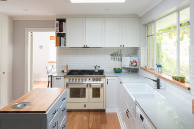 Inspiration for a transitional l-shaped separate kitchen in Canberra - Queanbeyan with a farmhouse sink, shaker cabinets, white cabinets, white splashback, brick splashback, white appliances, medium hardwood floors, with island, brown floor and grey benchtop.