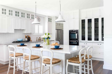 Photo of a large beach style u-shaped open plan kitchen in Perth with a farmhouse sink, raised-panel cabinets, white cabinets, marble benchtops, white splashback, subway tile splashback, black appliances, dark hardwood floors, with island and grey benchtop.