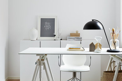 Minimalistisk inredning av ett mellanstort hemmabibliotek, med vita väggar, ljust trägolv och ett fristående skrivbord