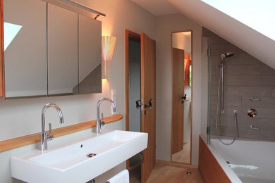 Modernes Badezimmer mit hellen Holzschränken, Einbaubadewanne, Duschbadewanne, Wandtoilette, Wandwaschbecken und hellem Holzboden in Sonstige