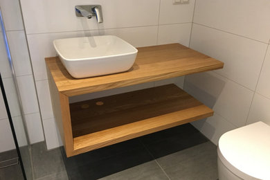 Modernes Badezimmer mit hellbraunen Holzschränken, Wandtoilette, weißen Fliesen, Aufsatzwaschbecken, Waschtisch aus Holz und grauem Boden in Frankfurt am Main