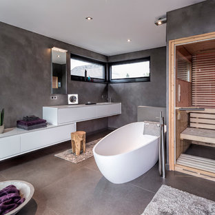 Mittelgroßes Modernes Badezimmer mit flächenbündigen Schrankfronten, weißen Schränken, freistehender Badewanne, bodengleicher Dusche, Wandtoilette, grauen Fliesen, grauer Wandfarbe, Sauna, grauem Boden, Betonboden, Unterbauwaschbecken und Falttür-Duschabtrennung in Düsseldorf