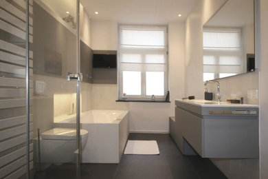 Diseño de cuarto de baño contemporáneo grande con armarios con paneles lisos, puertas de armario grises, bañera encastrada, sanitario de pared, baldosas y/o azulejos blancos, baldosas y/o azulejos de cerámica, paredes blancas, lavabo integrado y suelo gris