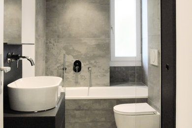 Cette image montre une petite salle de bain grise et blanche design avec un placard à porte plane, des portes de placard noires, une baignoire en alcôve, WC suspendus, un carrelage gris et une vasque.