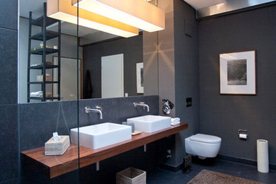 Ejemplo de cuarto de baño contemporáneo con baldosas y/o azulejos negros, encimera de madera, sanitario de pared, paredes grises, lavabo sobreencimera, bañera exenta, ducha abierta, encimeras marrones y espejo con luz