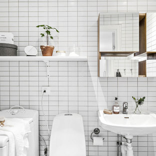 Foto på ett skandinaviskt badrum, med en toalettstol med hel cisternkåpa, vit kakel, vita väggar och ett väggmonterat handfat
