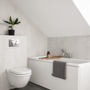 Minimalistisk inredning av ett litet badrum, med en vägghängd toalettstol, grå kakel, vit kakel, grått golv, ett platsbyggt badkar och vita väggar