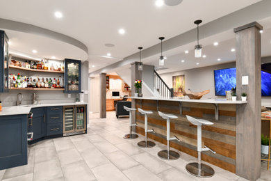 ワシントンD.C.にある広いトランジショナルスタイルのおしゃれな地下室 (ホームバー、グレーの壁、磁器タイルの床、標準型暖炉、レンガの暖炉まわり、グレーの床) の写真
