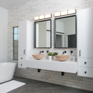 Modelo de cuarto de baño principal, industrial, de tamaño medio, con armarios con paneles lisos, puertas de armario blancas, bañera exenta, ducha a ras de suelo, sanitario de una pieza, baldosas y/o azulejos blancos, baldosas y/o azulejos de cerámica, paredes blancas, suelo de baldosas de cerámica, lavabo sobreencimera, encimera de cuarcita, suelo gris, ducha con puerta con bisagras y encimeras blancas