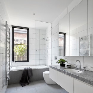 Photo of a large contemporary bathroom in Melbourne with white cabinets, a drop-in tub, a shower/bathtub combo, a wall-mount toilet, white tile, porcelain tile, white walls, porcelain floors, an undermount sink, marble benchtops, grey floor, an open shower, grey benchtops and flat-panel cabinets.