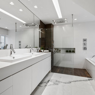 This is an example of a large contemporary ensuite bathroom in West Midlands with white cabinets, a wall mounted toilet, white tiles, white walls, an open shower, white worktops, flat-panel cabinets, a built-in bath, a walk-in shower, a built-in sink and white floors.