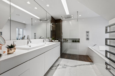 This is an example of a large contemporary ensuite bathroom in West Midlands with white cabinets, a wall mounted toilet, white tiles, white walls, an open shower, white worktops, flat-panel cabinets, a built-in bath, a walk-in shower, a built-in sink and white floors.