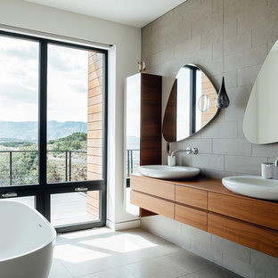 Imagen de cuarto de baño principal y doble, contemporáneo, de tamaño medio, con armarios con paneles lisos, puertas de armario de madera oscura, bañera exenta, baldosas y/o azulejos grises, baldosas y/o azulejos de cemento, paredes blancas, suelo de baldosas de porcelana, lavabo sobreencimera, encimera de madera, suelo gris y encimeras marrones
