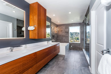 Exemple d'une douche en alcôve principale tendance en bois brun avec un placard à porte plane, une baignoire d'angle, un carrelage gris, un mur gris, un lavabo encastré, un sol gris, une cabine de douche à porte battante et un plan de toilette blanc.