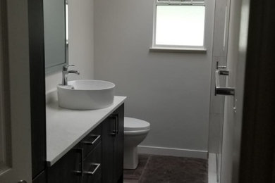 Example of a mid-sized beach style master brown tile and ceramic tile vinyl floor and brown floor alcove shower design in Vancouver with flat-panel cabinets, brown cabinets, a one-piece toilet, gray walls, a drop-in sink, quartzite countertops, a hinged shower door and white countertops