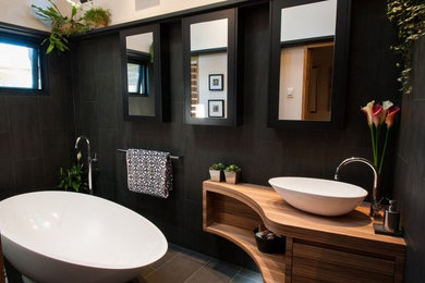 Modern inredning av ett mellanstort en-suite badrum, med möbel-liknande, skåp i mellenmörkt trä, ett fristående badkar, en dusch i en alkov, en toalettstol med hel cisternkåpa, svart kakel, porslinskakel, beige väggar, klinkergolv i porslin, träbänkskiva, svart golv, med dusch som är öppen och ett fristående handfat
