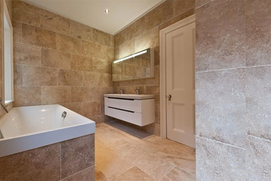 This is an example of a large contemporary family bathroom in London with freestanding cabinets, white cabinets, an alcove bath, beige tiles, stone tiles, white walls, limestone flooring, a built-in sink and beige floors.