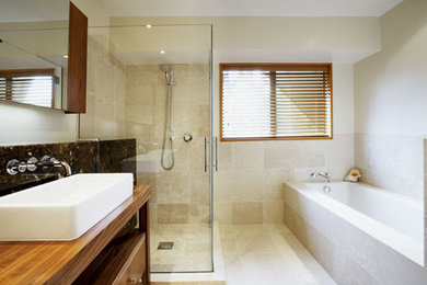 Modern inredning av ett badrum, med möbel-liknande, skåp i mellenmörkt trä, ett platsbyggt badkar, en toalettstol med hel cisternkåpa, stenkakel, beige väggar, marmorgolv, ett fristående handfat och träbänkskiva
