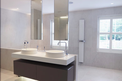 Inspiration for a large contemporary ensuite bathroom in Surrey with a console sink, solid surface worktops, an open shower, glass-front cabinets, a built-in bath, a walk-in shower, a wall mounted toilet, brown cabinets, grey tiles, porcelain tiles, grey walls, ceramic flooring and grey floors.