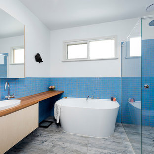 Inspiration for a large beach style master bathroom in Melbourne with flat-panel cabinets, light wood cabinets, wood benchtops, brown benchtops, a freestanding tub, a curbless shower, blue tile, white walls, a vessel sink and grey floor.