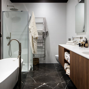Modelo de cuarto de baño principal, actual, grande, con armarios con paneles lisos, puertas de armario de madera oscura, bañera exenta, ducha esquinera, paredes blancas, suelo de baldosas de porcelana, lavabo integrado, suelo negro, ducha con puerta con bisagras y encimeras blancas