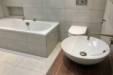 This is an example of a medium sized contemporary ensuite wet room bathroom in Kent with flat-panel cabinets, a built-in bath, a wall mounted toilet, grey tiles, porcelain tiles, grey walls, porcelain flooring, a vessel sink, wooden worktops and grey floors.