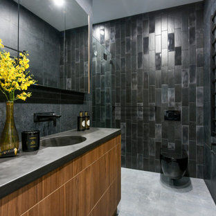 This is an example of a contemporary bathroom in Sydney with flat-panel cabinets, medium wood cabinets, a wall-mount toilet, black tile, an integrated sink, grey floor and grey benchtops.