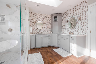 Cette photo montre une grande salle de bain principale tendance avec des portes de placard grises, une baignoire indépendante, un carrelage gris, un mur gris, un sol en bois brun, un sol marron, un plan de toilette blanc, des toilettes cachées, meuble double vasque et meuble-lavabo encastré.