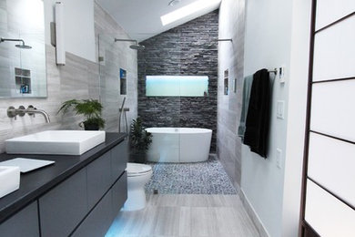 This is an example of a medium sized contemporary ensuite bathroom in Atlanta with flat-panel cabinets, grey cabinets, a freestanding bath, a built-in shower, grey tiles, stone tiles, limestone flooring, white walls, a two-piece toilet, a vessel sink, granite worktops, grey floors, an open shower, black worktops, a wall niche, double sinks, a floating vanity unit and a vaulted ceiling.