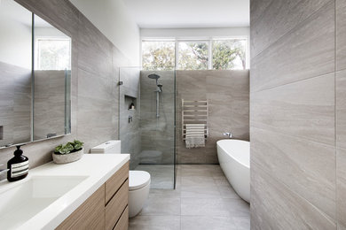 Idée de décoration pour une grande salle de bain principale design en bois clair avec une baignoire indépendante, une douche à l'italienne, WC à poser, un carrelage gris, des carreaux de porcelaine, un mur blanc, un sol en carrelage de porcelaine, un lavabo encastré, un plan de toilette en quartz modifié, un sol gris, une cabine de douche à porte battante, un plan de toilette blanc et un placard à porte plane.