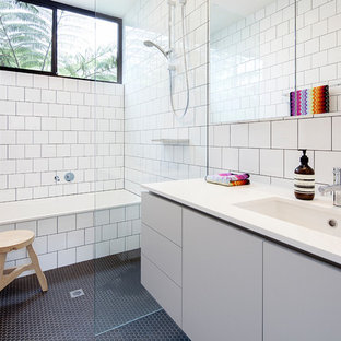 This is an example of a midcentury 3/4 bathroom in Melbourne with flat-panel cabinets, grey cabinets, a drop-in tub, a curbless shower, white tile, white walls, mosaic tile floors, an undermount sink, black floor, an open shower and white benchtops.