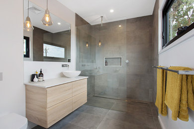 Modern inredning av ett badrum, med släta luckor, skåp i ljust trä, en kantlös dusch, grå kakel, vita väggar, ett fristående handfat, grått golv och med dusch som är öppen
