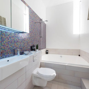 Medium sized contemporary ensuite bathroom in London with a built-in bath, a shower/bath combination, a wall mounted toilet, mosaic tiles, white walls, multi-coloured tiles, a wall-mounted sink and beige floors.