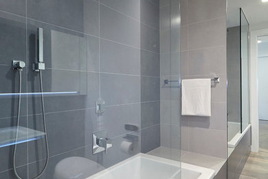 Inredning av ett modernt stort en-suite badrum, med ett platsbyggt badkar, en dusch/badkar-kombination, mellanmörkt trägolv, en toalettstol med hel cisternkåpa, grå kakel, porslinskakel, släta luckor, vita skåp, vita väggar, ett integrerad handfat, bänkskiva i glas och beiget golv
