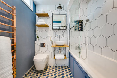 ロンドンにあるトランジショナルスタイルのおしゃれな浴室 (オープンシェルフ、ドロップイン型浴槽、シャワー付き浴槽	、壁掛け式トイレ、白いタイル、青い壁、ベッセル式洗面器、木製洗面台、マルチカラーの床) の写真