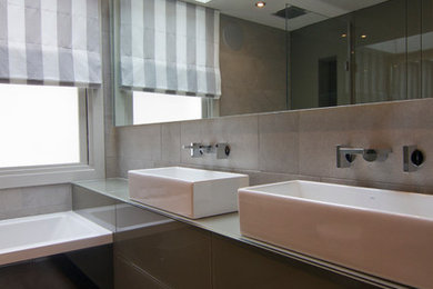 This is an example of a small contemporary bathroom in London with a console sink, glass-front cabinets, glass worktops, a built-in bath, a shower/bath combination, a wall mounted toilet, grey tiles, porcelain tiles, grey walls and porcelain flooring.