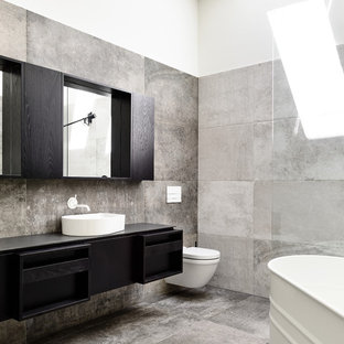 Inspiration for a mid-sized contemporary 3/4 bathroom in Melbourne with a freestanding tub, a wall-mount toilet, gray tile, ceramic tile, ceramic floors, grey floor, tile benchtops, black benchtops, flat-panel cabinets, black cabinets, white walls and a vessel sink.