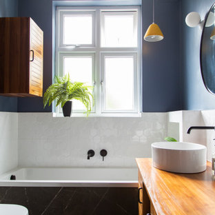 This is an example of a small transitional master wet room bathroom in London with louvered cabinets, brown cabinets, a drop-in tub, a wall-mount toilet, white tile, porcelain tile, blue walls, porcelain floors, wood benchtops, black floor, brown benchtops, a single vanity, a freestanding vanity and wood.