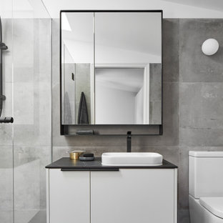 This is an example of a mid-sized contemporary 3/4 bathroom in Melbourne with flat-panel cabinets, a one-piece toilet, gray tile, ceramic tile, grey walls, ceramic floors, a vessel sink, solid surface benchtops, grey floor, a hinged shower door, black benchtops, a single vanity, a floating vanity, vaulted and white cabinets.