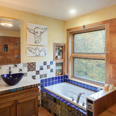 Example of an eclectic blue tile, multicolored tile and orange tile drop-in bathtub design in New York with recessed-panel cabinets, medium tone wood cabinets, yellow walls, a vessel sink and tile countertops