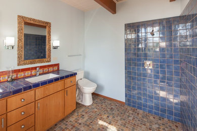 サンルイスオビスポにある広いサンタフェスタイルのおしゃれなバスルーム (浴槽なし) (フラットパネル扉のキャビネット、中間色木目調キャビネット、コーナー設置型シャワー、分離型トイレ、青いタイル、セラミックタイル、白い壁、トラバーチンの床、アンダーカウンター洗面器、タイルの洗面台、マルチカラーの床、オープンシャワー) の写真