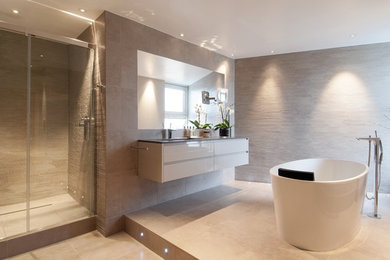 Photo of a large modern ensuite bathroom in London with a wall-mounted sink, a freestanding bath, a wall mounted toilet, porcelain tiles, porcelain flooring, flat-panel cabinets, beige cabinets, solid surface worktops, an alcove shower, beige tiles and beige walls.