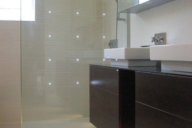 Inspiration for a large modern ensuite bathroom in London with flat-panel cabinets, dark wood cabinets, glass worktops, a corner bath, a walk-in shower, a wall mounted toilet, beige tiles, ceramic tiles, beige walls and ceramic flooring.