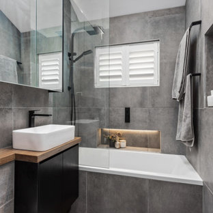Inspiration for a small contemporary master bathroom in Sydney with flat-panel cabinets, black cabinets, a drop-in tub, a shower/bathtub combo, a wall-mount toilet, gray tile, ceramic tile, grey walls, ceramic floors, a vessel sink, wood benchtops, grey floor, brown benchtops, a niche, a single vanity, a built-in vanity and vaulted.