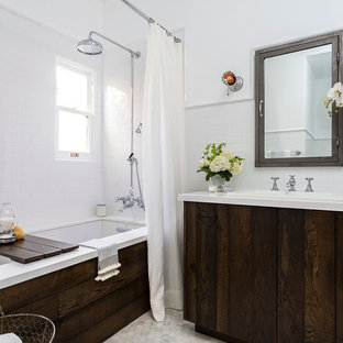 Inredning av ett lantligt litet en-suite badrum, med ett nedsänkt handfat, släta luckor, ett undermonterat badkar, en dusch/badkar-kombination, en toalettstol med separat cisternkåpa, vit kakel, tunnelbanekakel, vita väggar, marmorgolv, bänkskiva i akrylsten, skåp i slitet trä och dusch med duschdraperi