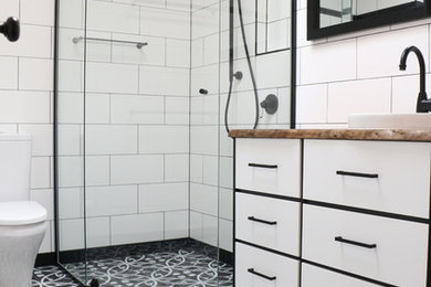 Inredning av ett 60 tals mellanstort badrum med dusch, med släta luckor, svarta skåp, en hörndusch, vit kakel, tunnelbanekakel, vita väggar, ett nedsänkt handfat, träbänkskiva och dusch med gångjärnsdörr