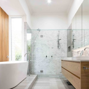 This is an example of a contemporary master bathroom in Melbourne with light wood cabinets, marble, white walls, cement tiles, engineered quartz benchtops, grey floor, flat-panel cabinets, a freestanding tub, a curbless shower, gray tile, a vessel sink, a hinged shower door and white benchtops.