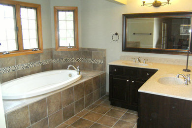 Example of a mid-sized classic master beige tile and ceramic tile ceramic tile and brown floor drop-in bathtub design in Indianapolis with raised-panel cabinets, brown cabinets, gray walls, an integrated sink and solid surface countertops